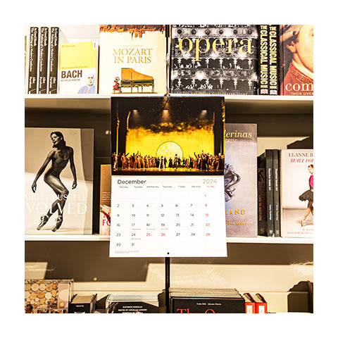 Royal Opera calendar open to December hanging on a shelf that is filled with books.