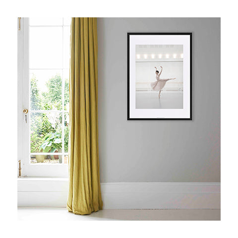 A print of a dancer framed and hanging on a grey wall beside a window.
