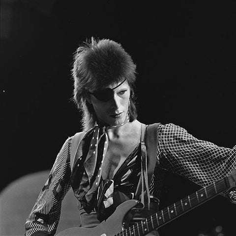 A black and white image of David Bowie wearing a polka dot neckerchief, patterned shirt and eyepatch.