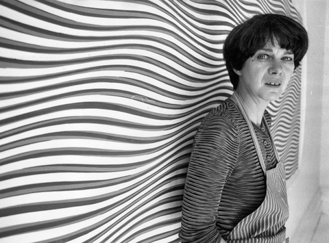 The artist Bridget Riley wears dungarees and a striped top. She stands in front of a large optical illusion painting featuring graphic, wavy lines.