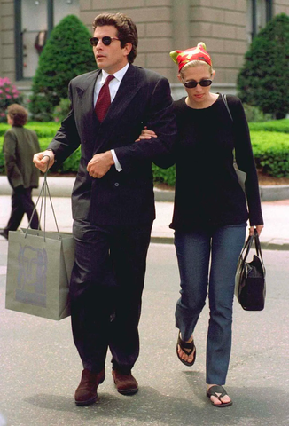 John F. Kennedy Jr. and wife Carolyn Bassette-Kennedy walk arm-in-arm through the city. He wears a suit, she is casually dressed in jeans, flip flops and sunglasses.
