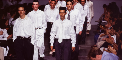 A group of male models, wearing oversized white shirts and black or white trousers, walk down a catwalk for the designer Commes des Garcons in 1995.