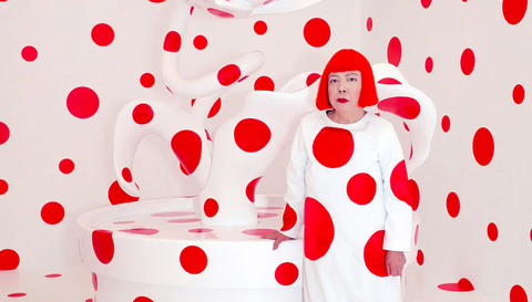 The artist Yayoi Kusama stands in front of one of her artworks. The art work is an abstract sculpture, white in colour featuring large red spots. The room they are in, and Kusama's dress both feature the same pattern. Kusama wears a short, bobbed wig that is bright red.