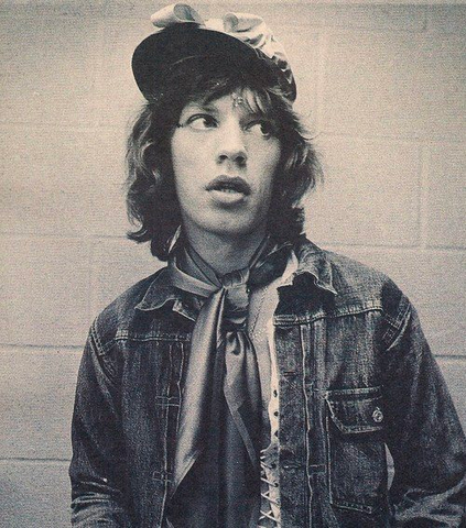 A black and white image of a young Mick Jagger, dressed in a denim jacket and silk neckerchief, stands in front of a brick wall.