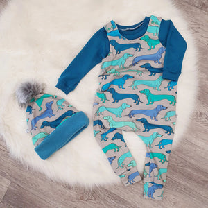 bayridgecaskandkeg sausage dog printed dungaree romper. The romper has a grey background with blue and green miniature dachshunds. The romper is paired with a teal lonsleeve t-shirt and matching dachshund print bobble hat with teal fleece lining and fluffy grey pom