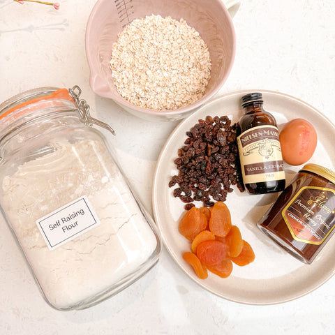 Image of ingredients laid down on a work surface incusing cinnamon honey, oats, flour, egg, raisins and dried apricot  