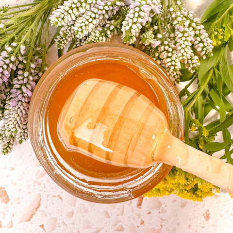 Photo of the top of a jar of honey surrounded by purple flowers with green leaves with a honey spoon