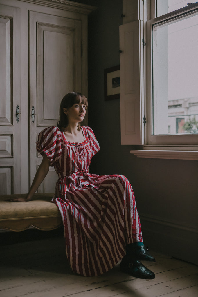 Rosie sits on the bed wearing Sadie Dress from Little Tienda's Picnic Collection