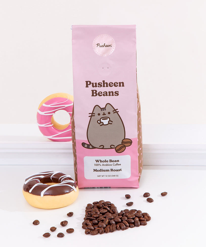 Pusheen Beans Coffee bag sits on white surface. Coffee bag shows Pusheen the Cat holding a cup of coffee on the front of the bag with additional coffee information. Coffee bag is surrounded by coffee beans in the shape of a heart and glazed donuts.