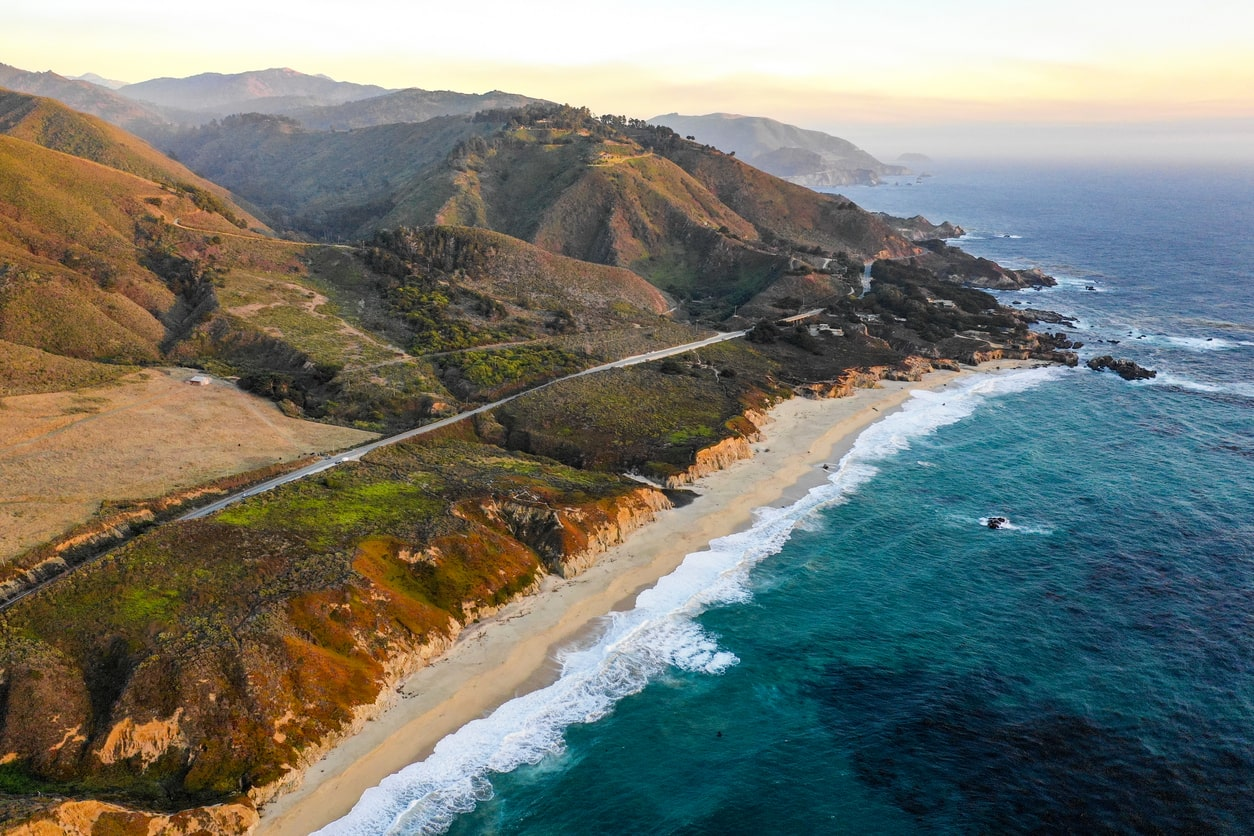 Aerial view of a coastal road winding through the rugged landscape of Big Sur, featuring cliffs, beaches, and mountains during sunset | KEUTEK