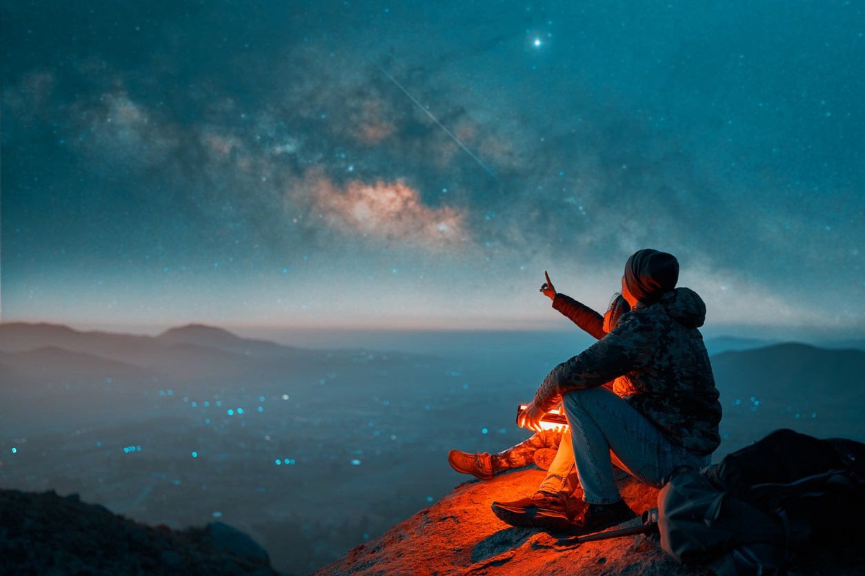 Camper sitting on a mountain edge at dusk, pointing at a shooting star in a clear night sky, representing the adventure and essentials of novice boondocking | KEUTEK