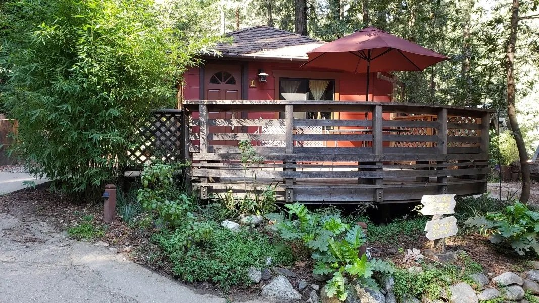 Cozy red cabin with a spacious wooden deck and a red umbrella, nestled among lush greenery in Riverside Campground & Cabins, creating a peaceful retreat in a natural setting | KEUTEK