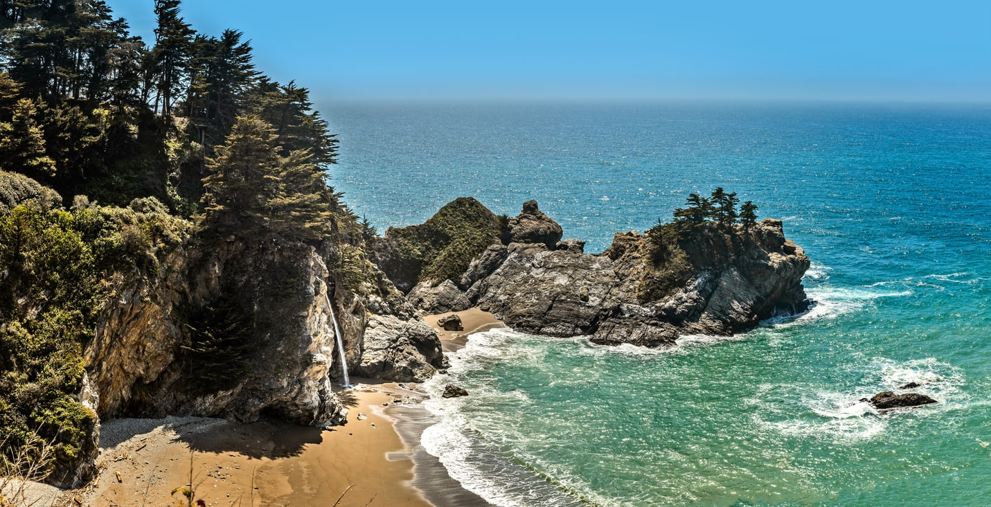 Panoramic view of a secluded cove in Pfeiffer Big Sur State Park, featuring rugged cliffs, dense evergreen trees, and a small waterfall cascading into a turquoise sea | KEUTEK
