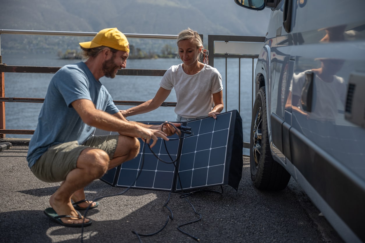 Two campers setting up a portable solar panel next to their van by a lake, harnessing renewable energy for their wilderness experience | KEUTEK