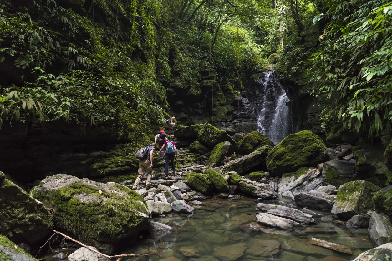 Hikers navigating a rocky stream in a lush forest with a waterfall in the background, symbolizing exploration in public lands and forests | KEUTEK