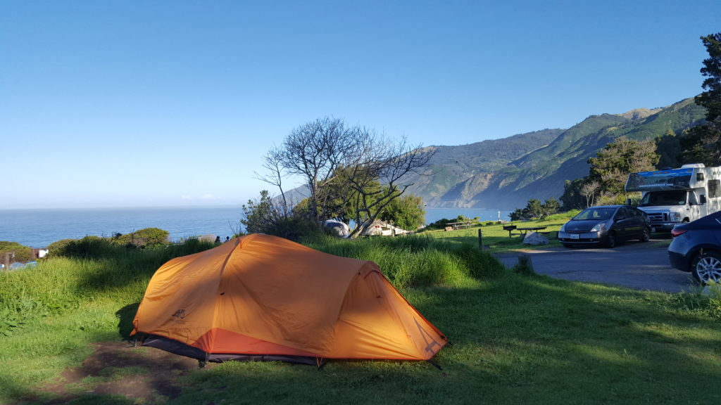 Scenic camping site at Kirk Creek Campground with a view of the Pacific Ocean and steep coastal mountains, featuring an orange tent, vehicles, and RVs under a clear blue sky | KEUTEK