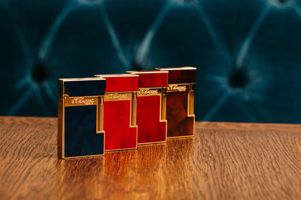 Four S.T. Dupont Ligne 2 lighters, in blue, red, and brown, with gold finishes, displayed on a wooden surface against a blue textured background