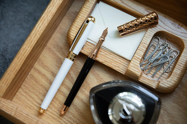 An elegant desk setup featuring a white and gold S.T. Line D fountain pen, a black S.T. Dupont Liberte fountain pen with gold accents, a carved golden cap, a paper note block, and paper clips in a wooden tray, with a glass inkwell in the foreground.