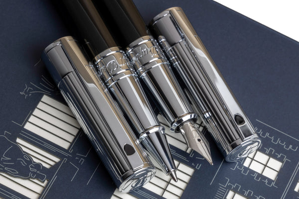 A close-up image of two black and chrome S.T. Dupont D-Initial pens, including a fountain pen, and rollerball, arranged diagonally on a blue background with schematic designs