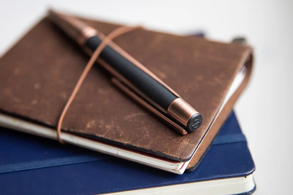 A premium black S.T. Dupont Defi ballpoint pen with copper details, secured by a leather band on a worn brown leather journal.