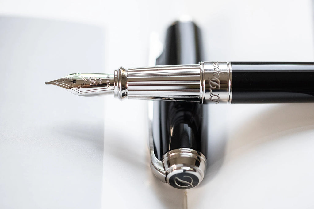 A close-up photograph of a luxury S.T. Dupont fountain pen with a detailed grip section. The pen features a black lacquer barrel and a palladium cap with a clip. The nib is crafted from 14K gold and is intricately designed with an S.T. dupont logo