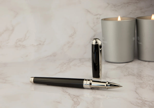A luxury S.T. Dupont black Line D rollerball pen with palladium accents, resting on a marble surface next to a pair of silver candles in cylindrical holders.