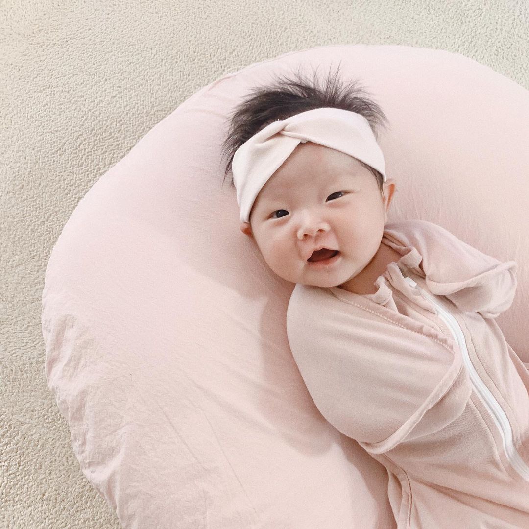 Smiling baby in her pink swaddle lying on a baby pink lounger. She’s wearing a light pink headband. Click to visit the baby pink swaddle product page.