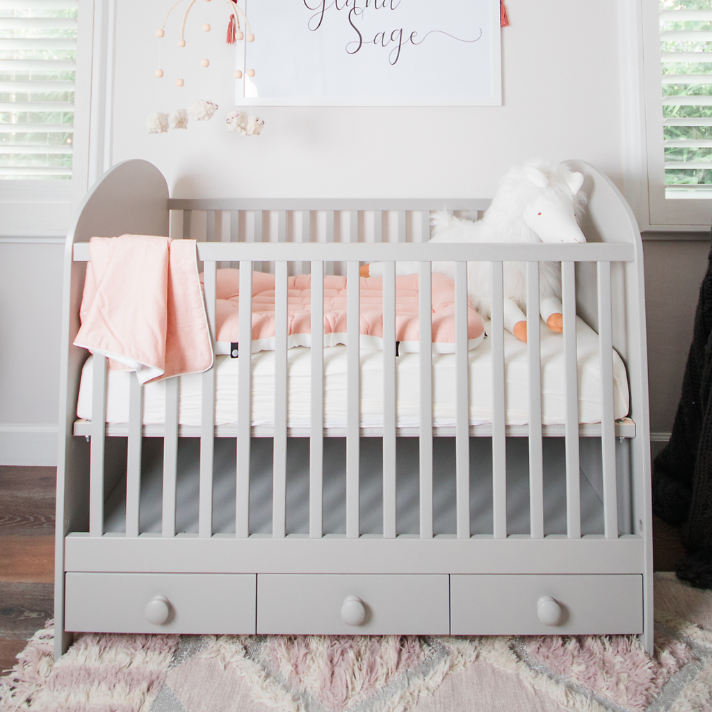 A crib painted in light gray which contains a baby pink relaxer and white stuffed hourse. A baby pink double blanket hangs on the crib. The crib is in a nursery with white paint.