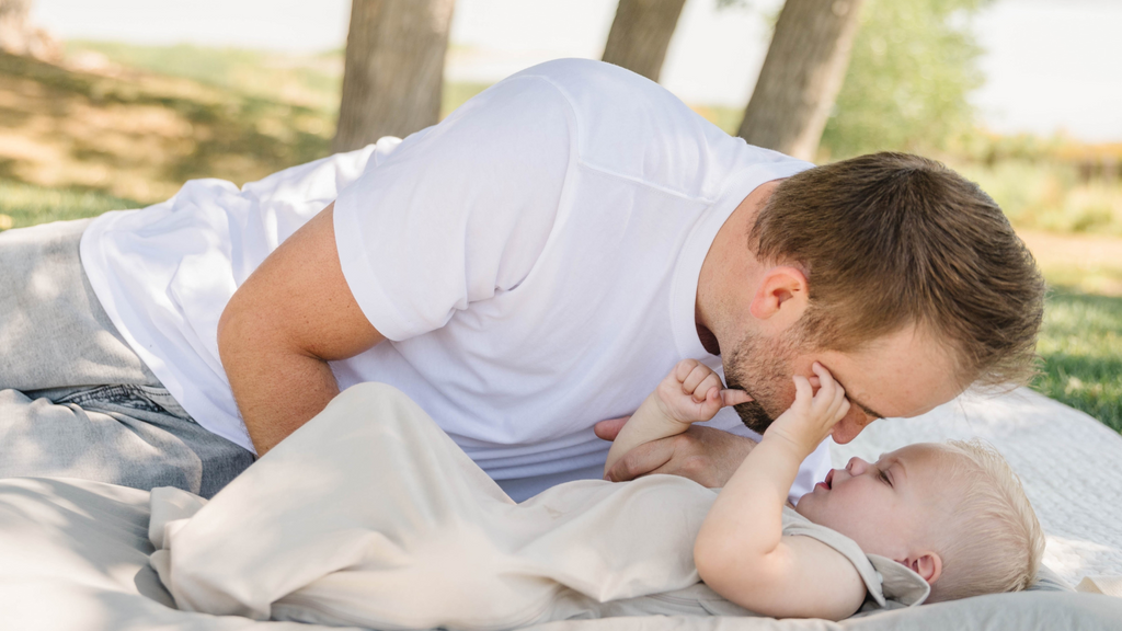 A father and a baby in his oat beige sleep sack having a picnic in the park. The father's head is close to the baby's. He seems to be hugging him.