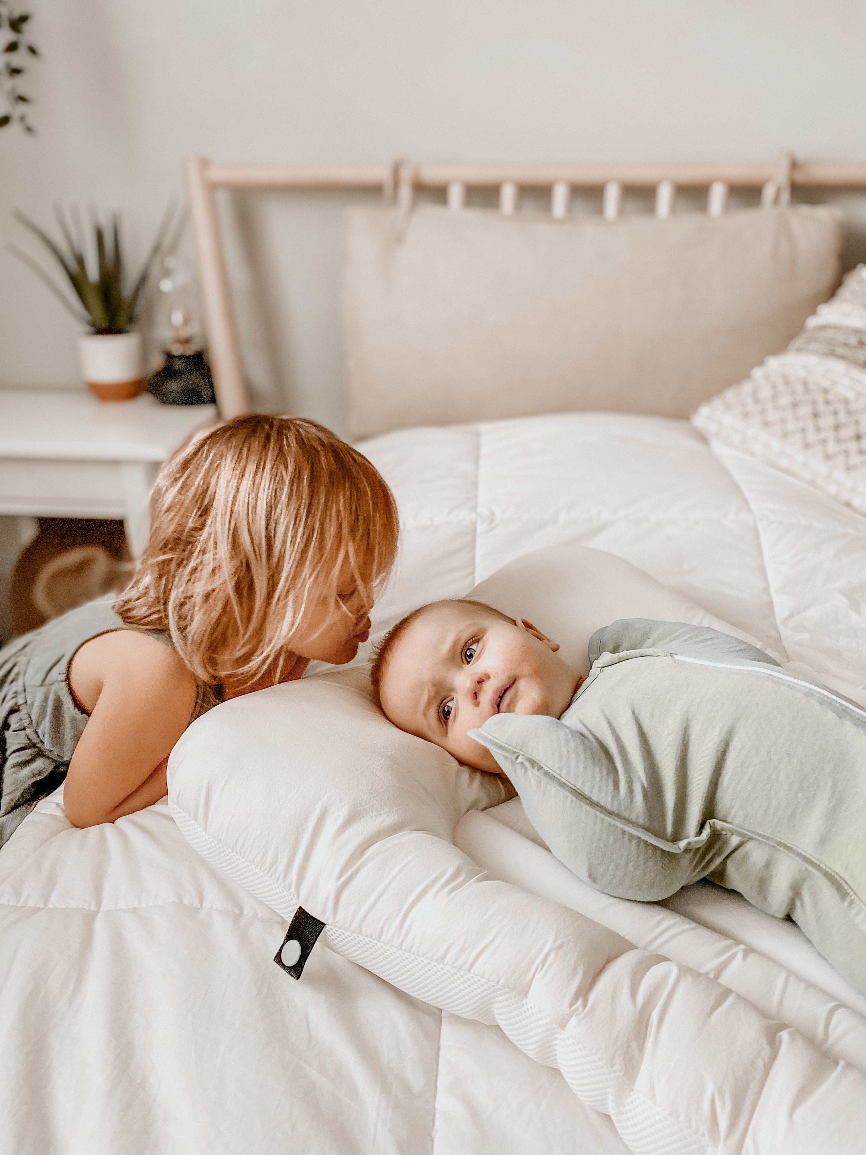 A little girl kissing her baby brother. The brother is wearing a swaddle and lying on a baby mattress or relaxer.