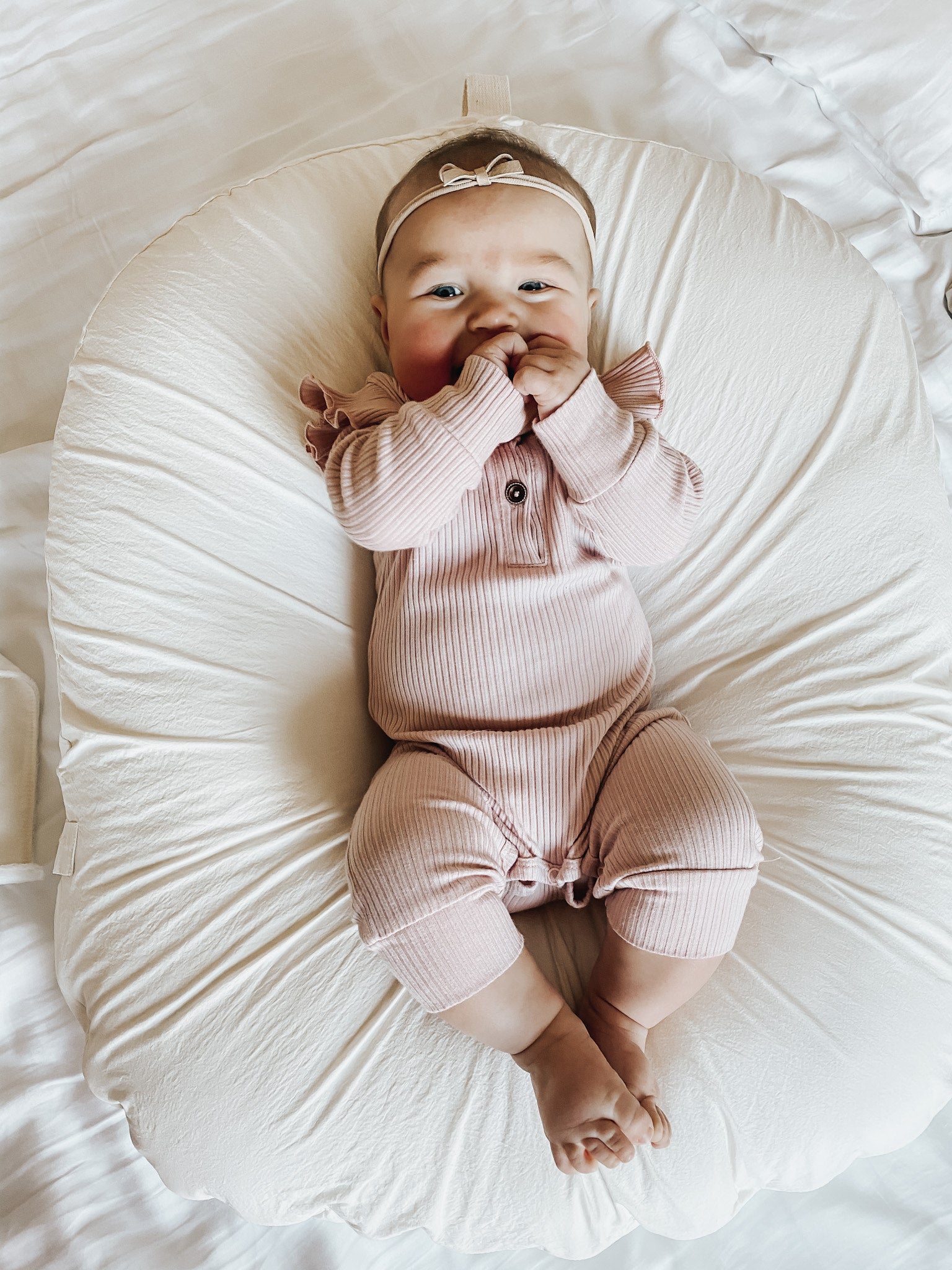 A baby facing the camera, holding both of her hands to her mouth. She’s wearing a light pink onesie and has a cute light pink ribbon headband. She has her feet together and is lying on a cream baby lounger.