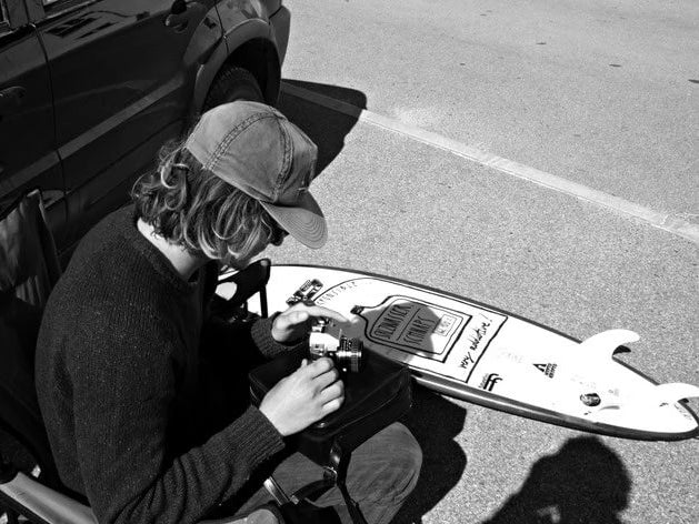 Laurenz Steierl probiert seine analoge Kamera des Typs Minolta X700 aus. Neben ihm liegt ein Surfboard.