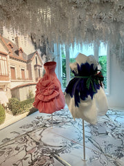 Two Dior dresses, the one on the left is deep rose coloured with ruffles, the one on the right with dark blue feathers. White fake flowers hanging down from the ceiling.