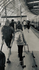 Black & White photo of a main and girl on a station platform, walking along with backpacks on, pulling wheelie cases.