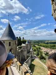 Colour photo looking out over the edge of a medieval walled city, with grey pointed turrets. There is blue sky and green trees.