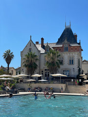 Colour photo of a white chateau, with a grey roof, and three palm trees in the front. In the foreground is a swimming pool.