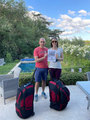 A white man, wearing a red tshirt and blue shorts, standing next to a white woman, wearing a white tshirt and burgundy leggings. They are holding a sheet of white paper. In the foreground are two red and black suitcases