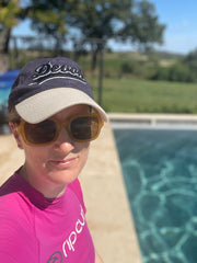 Colour photo of white female in her 40s, wearing a pink sun top, yellow sunglasses, and navy baseball cap with a beige peak, looking at the camera, with a swimming pool in the background