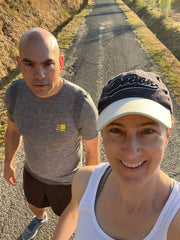 Colour photo of a bald white male in a grey stop and black shorts, standing slightly behind a white woman in white vest top and a navy peak cap.