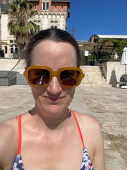 Colour photo of a white female in her 40s, looking at the camera. She is wearing a pair of yellow sunglasses and a blue and white bikini top with bright orange straps