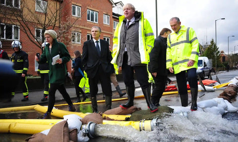 Ex-UK Prime Minister Boris Johnson, wearing a yellow high-vis jacket and welly boots, standing by large pipes on a road, with water gushing out |Contented Earth | Climate Confidence | Carbon Literacy Training