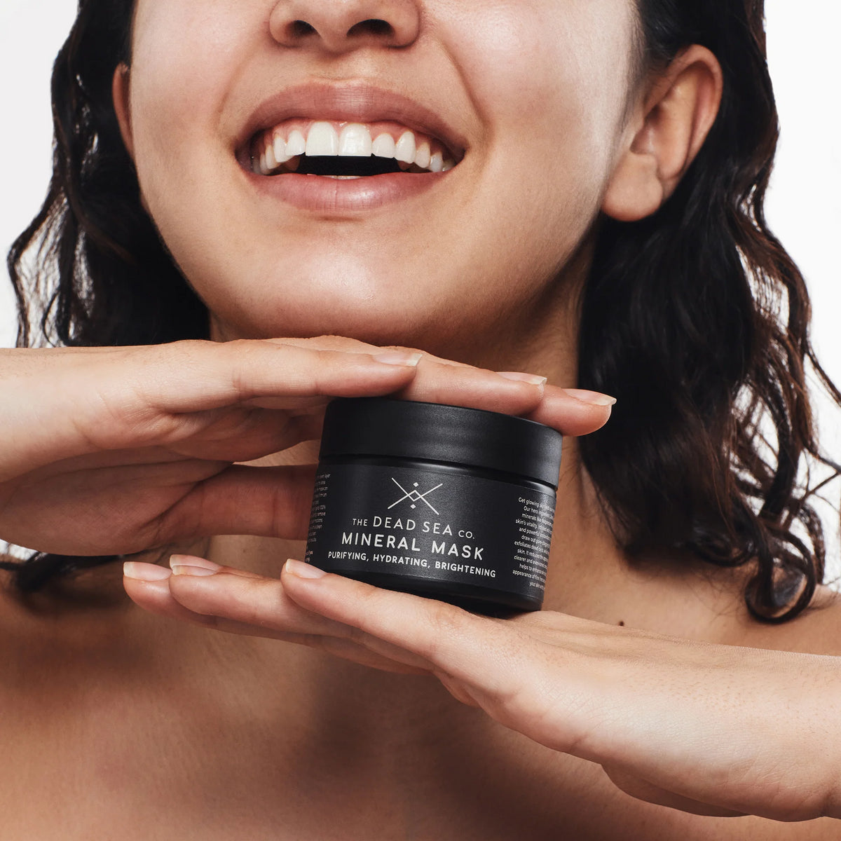 The image shows a woman with a bright smile and clear, dry skin holding The Dead Sea Co's Dead Sea mud mask between her hands. The focus is on the woman's smile and lower half of her face, which is clean and radiant, with no visible blemishes or imperfections as well as the black glass jar containing the Dead Sea mud mask. The packaging has a sleek and elegant appearance, with a minimalist design that exudes a sense of sophistication. The jar is reflective, and the black colour adds a touch of luxury to the overall aesthetic. The overall impression is one of confidence and self-care.