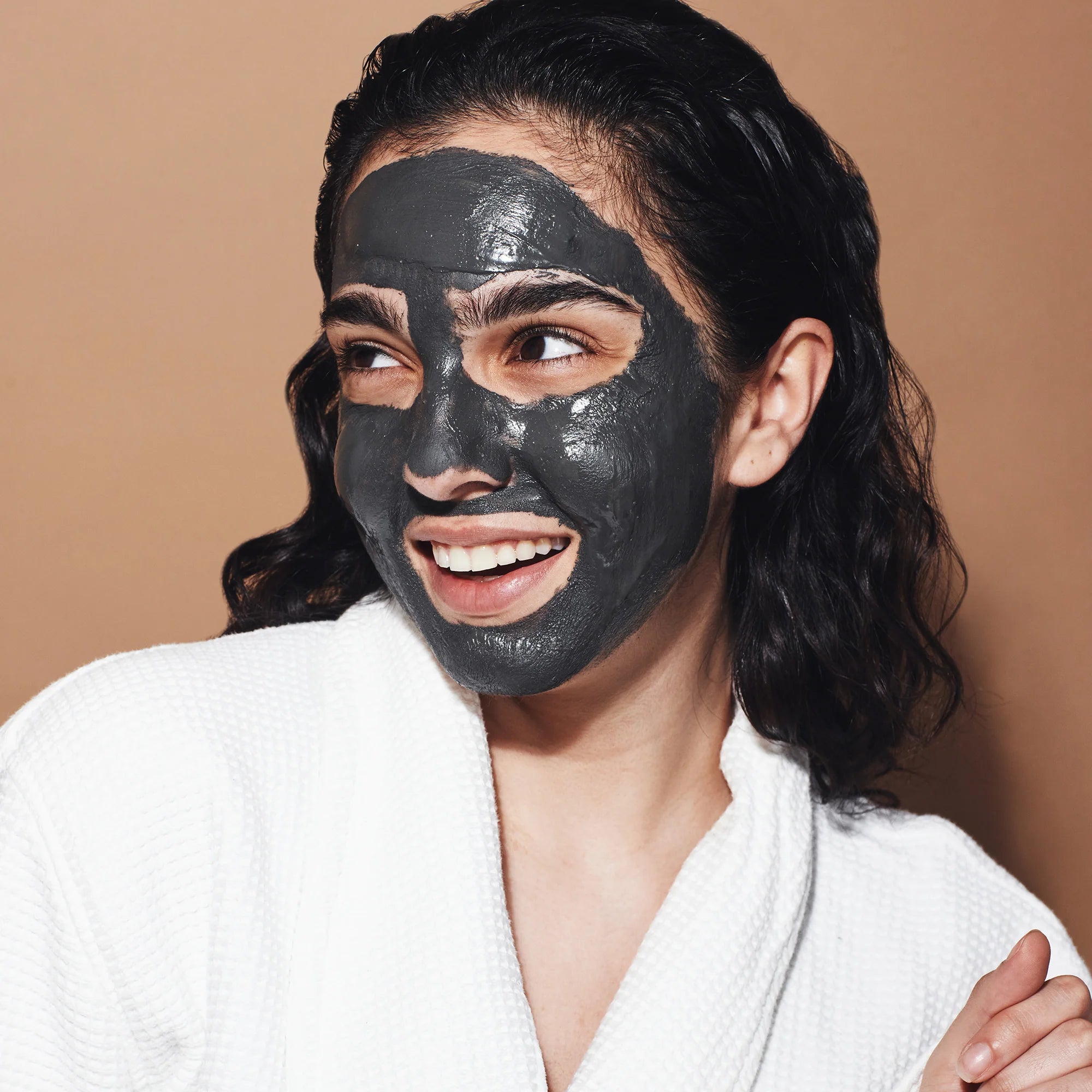 In the image, a woman smiles contentedly while indulging in a pampering session with a thick, creamy layer of dark grey Dead Sea mud mask covering her face. The mask's nourishing texture is visible as she looks away from the camera.