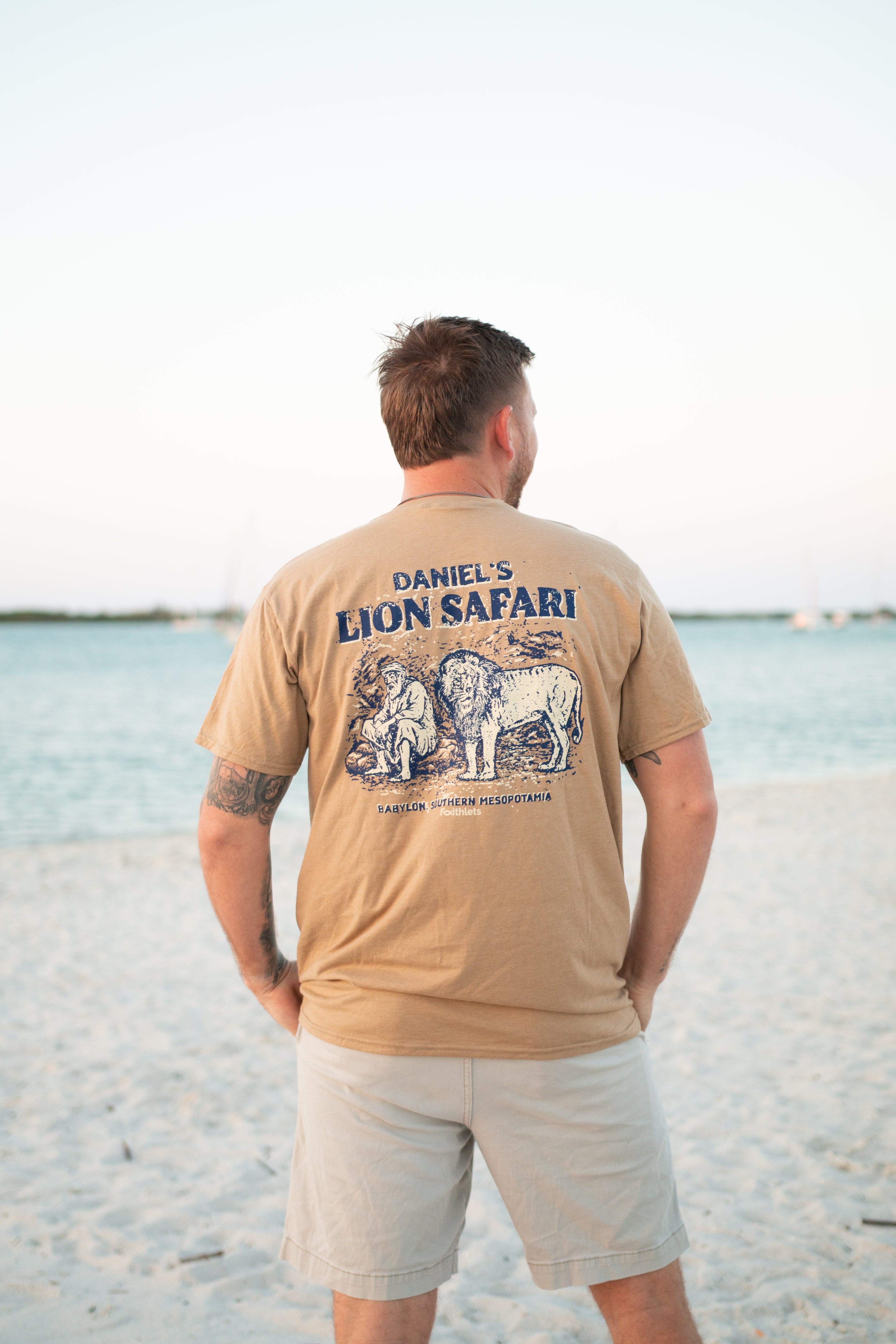 Man in a 'Daniel's Lion Safari' t-shirt standing on a beach, facing the ocean.