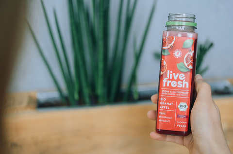 Hand hält eine Flasche 'live fresh' Bio-Granatapfel-Apfelsaft vor einer Zimmerpflanze.