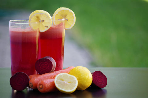 Zwei Gläser rote Beete Saft mit Zitronenscheiben dekoriert, daneben liegen rote Beete und Karotten.