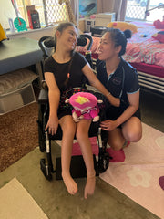 휠체어에 앉아 있는 엠마 디데(Emma Deede)는 옆에 웅크리고 있는 물리치료사 달레나 판나(Dalena Pangna)와 즐거운 순간을 나누고 있습니다. 두 사람 모두 서로를 향해 따뜻하게 미소를 짓고 있어 긍정적이고 격려적인 물리 치료 세션을 강조하고 있습니다.