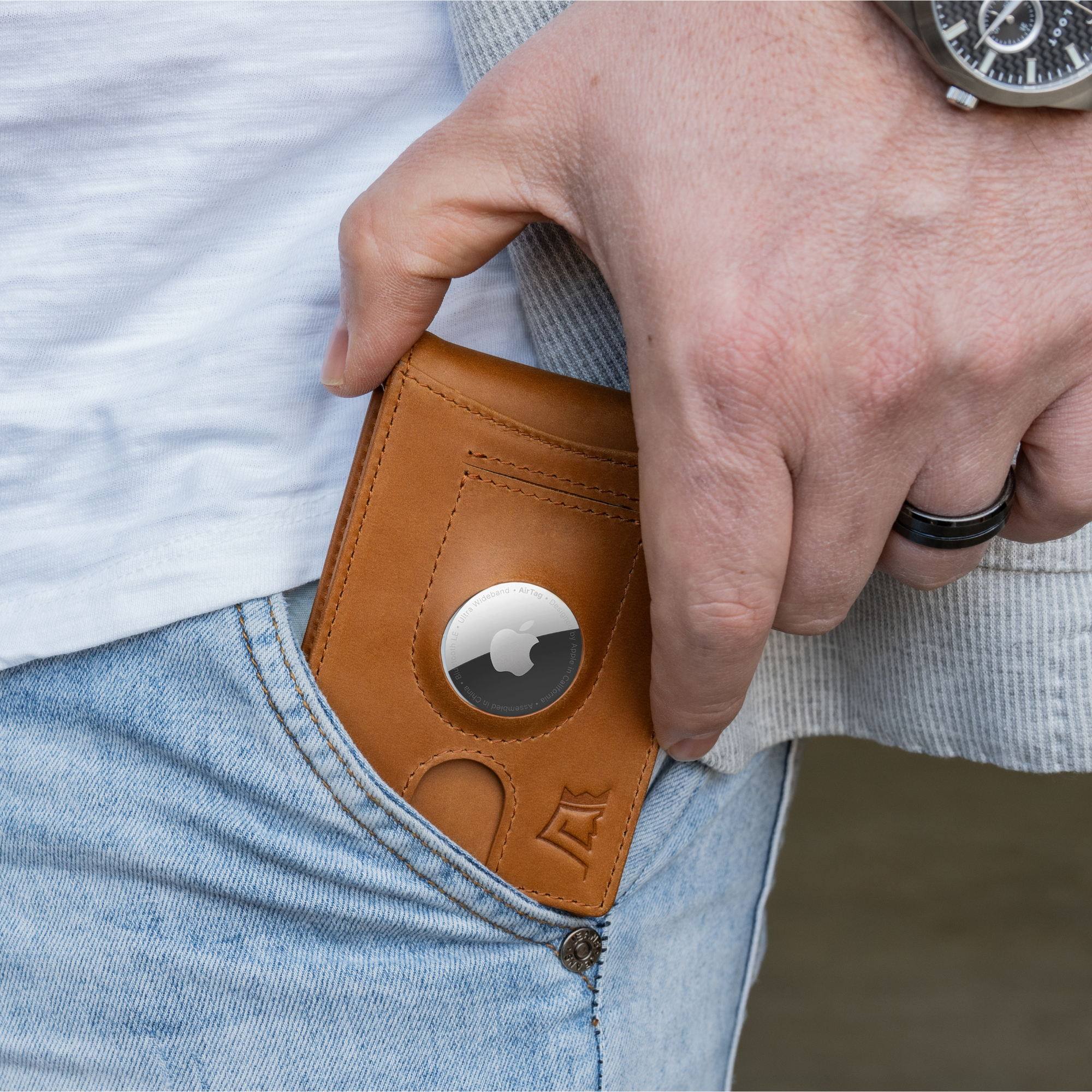Person inserting an Apple AirTag into a brown leather wallet in their front jeans pocket.