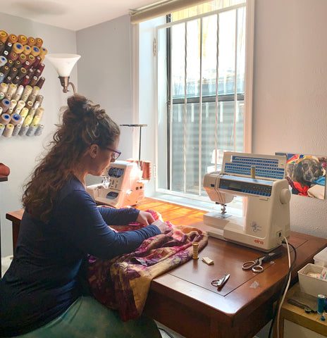 woman with long brown hair hand sewing a colorful jacket at sewing machine