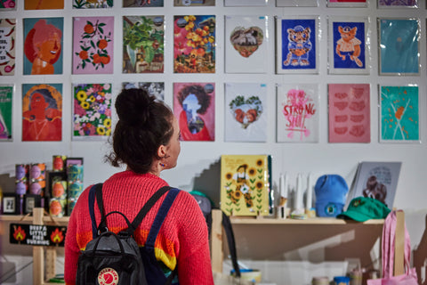 A young woman looking up at a wall inside a shop called Flock and Gaggle, on the wall is lots of independent artists work 